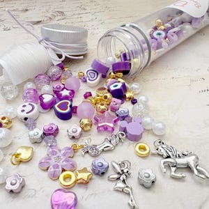 May include: A collection of purple, white, and gold beads and charms, including stars, flowers, hearts, and smiley faces, spilling from a clear tube. The beads are scattered on a light-colored surface, suggesting a crafting or jewelry-making theme.