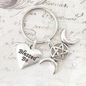 May include: Silver-coloured keychain with charms. The charms include a heart with "Blessed Be" text, a pentagram, and crescent moons. The keychain is on a silver ring.