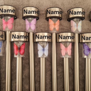 May include: Ten solar garden lights with silver metal stakes and clear cylindrical housings. Each light features a butterfly graphic in various colours including pink, purple, orange, blue, and white. The word Name is printed on each silver cap.