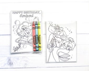 May include: Two rectangular coloring sheets featuring black and white line art of mermaids and a merman with a trident. The left sheet includes the text HAPPY BIRTHDAY, Amber! and is packaged with three Crayola crayons in red, blue, and yellow.