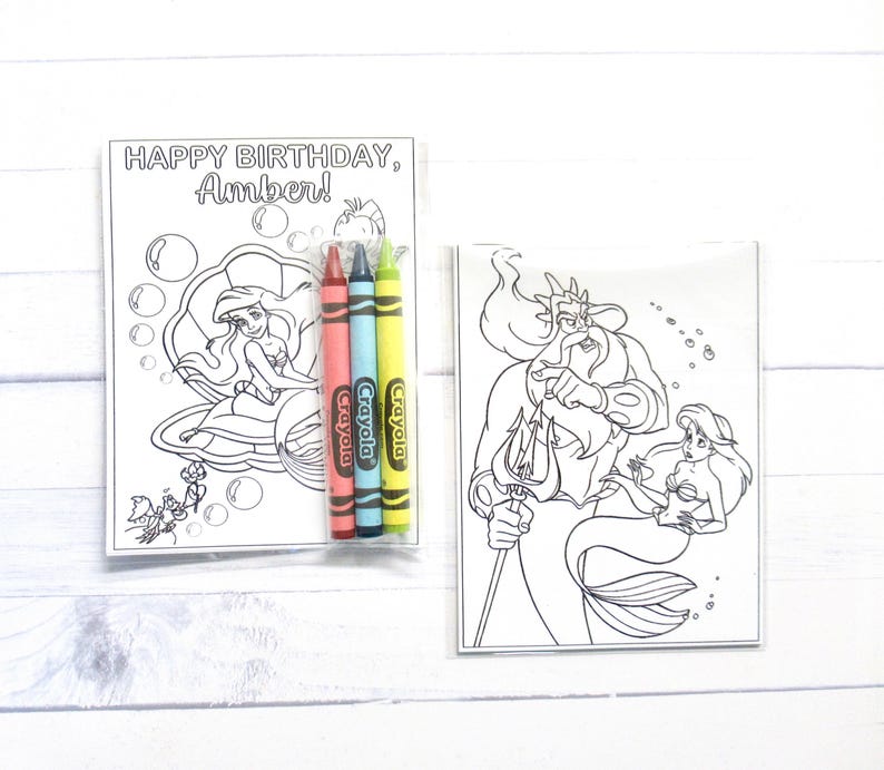 Puede incluir: Dos p&aacute;ginas para colorear en blanco y negro con Ariel y el Rey Trit&oacute;n de La Sirenita. Una p&aacute;gina tiene el texto "HAPPY BIRTHDAY, Amber!" con Ariel en una concha. Se incluyen tres crayones Crayola rojos, azules y amarillos.