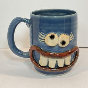 May include: A blue ceramic mug featuring a sculpted face with two protruding white eyes, dark pupils, and a wide brown mouth displaying three prominent white teeth. The mug has a curved handle on the left side and a smooth, glossy finish throughout.