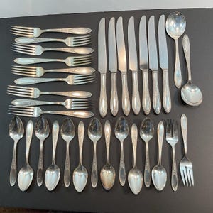 May include: A collection of vintage silverware, including forks, knives, spoons, and serving spoons, arranged on a dark surface. The silverware has a silver finish with ornate handle detailing. The arrangement showcases a complete set of dining utensils.