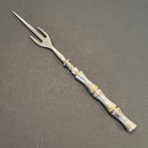 May include: A silver-toned metal cocktail fork featuring a three-pronged head with a long, pointed central tine. The handle is designed with a textured, segmented bamboo-like appearance, transitioning into a smooth, flared base at the end.