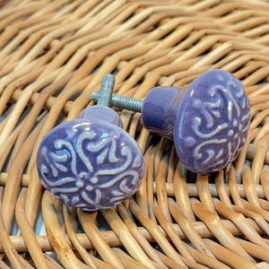 May include: Two round ceramic cabinet knobs in a deep purple hue, each featuring a raised, light-colored floral relief pattern. The knobs have a smooth, glossy finish and are attached to a silver-toned metal screw post for installation.