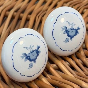 May include: Two round white ceramic cabinet knobs featuring a smooth, glossy finish. Each knob is decorated with a central blue floral pattern enclosed within a delicate, scalloped blue circular line, creating a clean and uniform appearance.