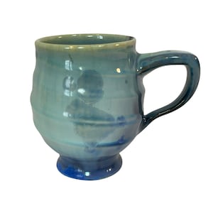 May include: A ceramic mug with a rounded body and a pedestal base, featuring a glossy glaze in shades of light blue, teal, and deep navy. The surface has a subtle horizontal ribbed texture and organic, cloud-like crystalline patterns.