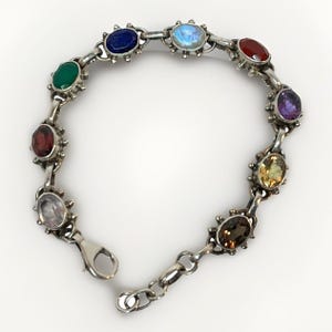 May include: Silver bracelet featuring a series of colorful gemstones, including red, green, blue, and purple stones. The bracelet has a lobster clasp closure and is designed with a series of oval-shaped stones.