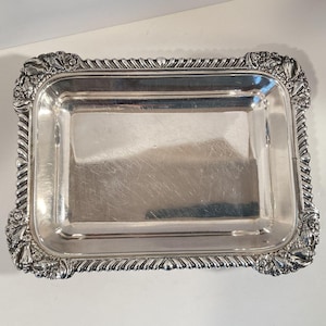 May include: A rectangular silver-colored tray with ornate floral and rope-like detailing around the edges. The tray has a reflective surface and is likely made of metal. The corners feature decorative floral designs.
