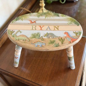 May include: A small, wooden step stool with an oval top. The top is painted with a dinosaur theme, including various dinosaurs, plants, and the name "RYAN". The legs are painted with a matching design.