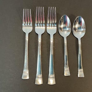 May include: A set of five silver-colored utensils: three forks and two spoons. The forks have four tines, and all utensils have decorative detailing on the handles. The silverware is arranged on a dark surface.
