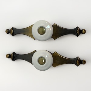 May include: Two cabinet pulls with a white ceramic center and a brass and dark bronze metal design. The pulls have a decorative shape with a round center and are set against a white background.