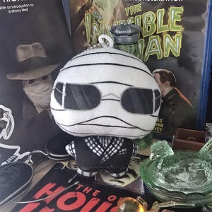 May include: A soft toy of the Invisible Man, wrapped in white bandages and wearing sunglasses and a black suit. The background features film and book covers related to the Invisible Man and The House of Horror. The toy is a keychain.
