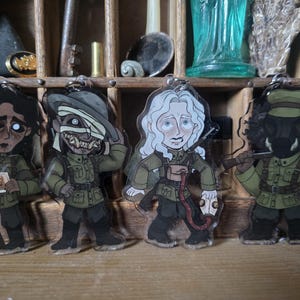 May include: Four clear acrylic charms featuring illustrated characters in olive green military uniforms. The characters include a person holding a small photograph, a figure with a bandaged face, a person with white hair, and a masked figure holding a rifle.