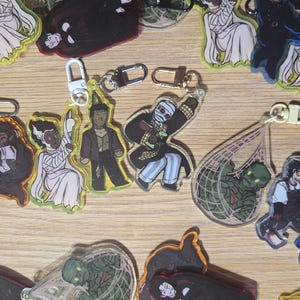 May include: A selection of acrylic keyrings with cartoon character designs. The keyrings have gold-coloured clasps and are displayed on a light brown wooden surface. The characters are in various colours, including black, white, blue, and green.