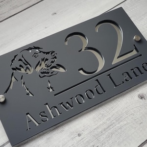 Può includere: Un cartello per numeri civici grigio scuro con il numero 32 in argento e il nome della strada Ashwood Lane. Il cartello presenta un design ritagliato a forma di testa di cane.