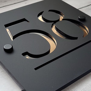Könnte beinhalten: Schwarze Hausnummer-Plakette mit der Nummer 58, die in einer modernen Schriftart ausgeschnitten ist. Die Nummer ist golden und die Plakette ist schwarz.