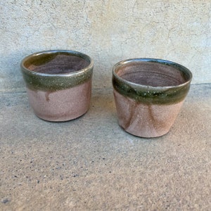 Puede incluir: Dos tazas de cerámica marrón con un glaseado verde alrededor de la parte superior. Las tazas están sobre una superficie de hormigón.