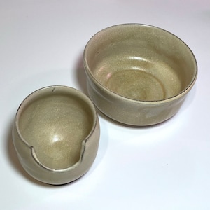 Op de afbeelding: Twee handgemaakte keramische kommen in een neutrale, aardse tint. Eén is een kleine, afgeronde kom met een gekerfde zijkant, de andere is een grotere, open kom. Beide hebben een matte afwerking en een eenvoudig, minimalistisch ontwerp.