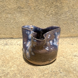 May include: A small, dark brown ceramic vase with a unique, irregular shape and a speckled glaze. The vase has a light blue and white speckled glaze with a brown base.