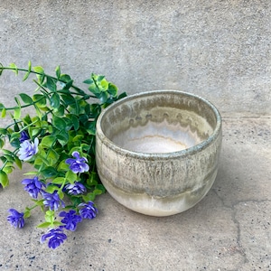 May include: A small, light green ceramic bowl with a white glaze on the inside. The bowl has a textured, speckled surface and is sitting on a gray concrete surface. A sprig of purple flowers is in the foreground.