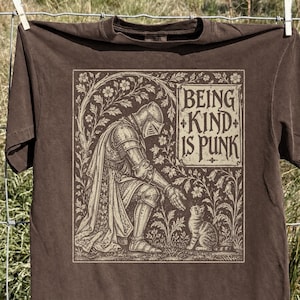 May include: A brown short-sleeved t-shirt featuring a graphic print of a knight in armor kneeling before a small cat. The illustration is framed by floral vines and includes the text "BEING KIND IS PUNK" in a decorative, stylized font.