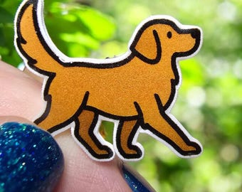 May include: A cartoon sticker of a golden retriever dog. The dog is depicted in a walking pose with a light brown body and a black outline. The sticker has a white border. The background is blurred green foliage.