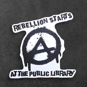 May include: A white sticker with black text and graphics. The text reads "REBELLION STARTS AT THE PUBLIC LIBRARY" above a black anarchy symbol. The sticker has a distressed, spray-painted aesthetic.