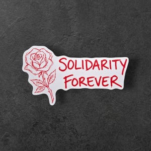 Pode incluir: Um autocolante branco com uma ilustração de rosa vermelha e as palavras "SOLIDARITY FOREVER" em vermelho. O autocolante está sobre uma superfície texturizada cinza escuro. A rosa é detalhada.