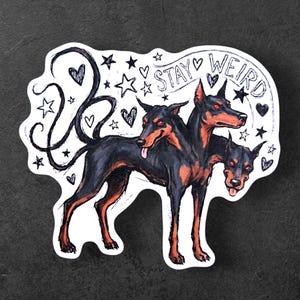 Puede incluir: Una pegatina con un perro de tres cabezas en negro y rojo, con el texto "STAY WEIRD" en un estilo manuscrito. El diseño incluye estrellas, corazones y espirales, creando una estética caprichosa y atrevida.