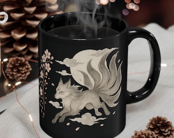 May include: Black ceramic mug featuring a watercolor illustration of a nine-tailed fox running across a moonlit sky. The design includes cherry blossoms and clouds. The mug has a curved handle and is filled with a steaming dark liquid.