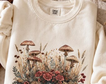 May include: A cream-colored crewneck sweatshirt featuring a Comfort Colors label at the inner neckline. The front displays a detailed graphic illustration of various brown mushrooms, tall dried grasses, and clusters of small red flowers.