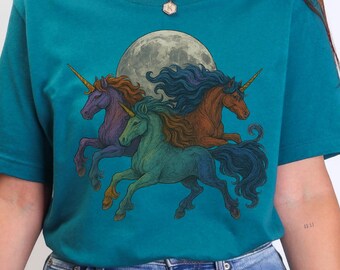 May include: Teal t-shirt featuring a graphic of three unicorns with colorful manes and horns, set against a full moon. The unicorns are in shades of purple, green, and brown. The shirt is made of a soft material.