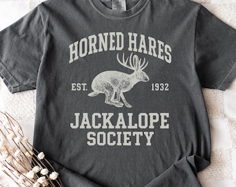 May include: A charcoal gray short-sleeve t-shirt featuring a cream-colored graphic of a jackalope. The text reads HORNED HARES, EST. 1932, and JACKALOPE SOCIETY in a collegiate font. The design is centered on the chest of the garment.