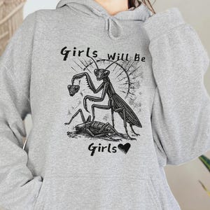 May include: A light gray hoodie featuring a black graphic of a praying mantis with the text "Girls will be Girls" in a handwritten font. The hoodie has a drawstring hood and a kangaroo pocket.