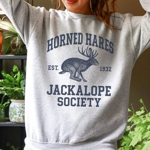 May include: A light gray crewneck sweatshirt featuring a dark blue graphic print. The design includes a central illustration of a jackalope with the text HORNED HARES, EST. 1932, and JACKALOPE SOCIETY printed in a bold, collegiate-style font.
