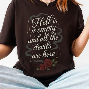 Puede incluir: Camiseta marrón oscuro con la cita "Hell is empty and all the devils are here" en escritura blanca. El diseño incluye un remolino ahumado y dos rosas rojas en la parte inferior. La camiseta la lleva una persona sentada con las piernas cruzadas.