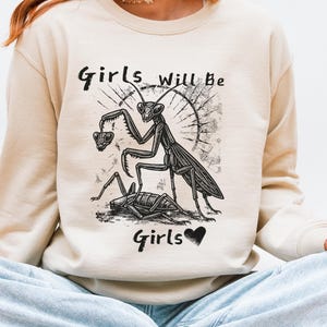 Pode incluir: Camisola creme com um gráfico preto de um louva-a-deus segurando um coração e o texto "Girls will be Girls" em uma fonte estilizada. O design inclui um símbolo de coração. A camisola é de estilo casual.