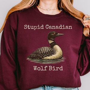 Puede incluir: Sudadera burdeos con el texto "Stupid Canadian" sobre una ilustración de un somorgujo, y las palabras "Wolf Bird" debajo. El somorgujo es negro, blanco y beige, representado en el agua.