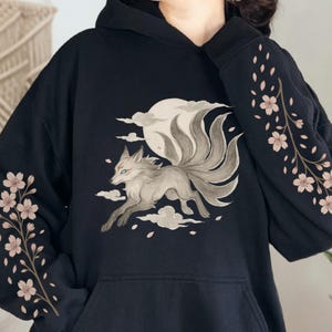 May include: Black hoodie with a nine-tailed fox design, full moon, and clouds. Floral branch design on the sleeves. The fox is grey and white with green eyes. The hoodie has a front pocket.