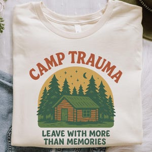 May include: Cream-colored t-shirt with the words "CAMP TRAUMA" in orange lettering above a graphic of a cabin in the woods. The text "LEAVE WITH MORE THAN MEMORIES" is below the cabin graphic.