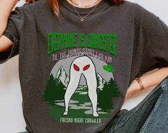 Op de afbeelding: Een donkergrijs T-shirt met een groene, witte en rode grafische print van een wezen met rode ogen en de tekst "Everyone is Gangster Til the Pants Start Walkin' Fresno Night Crawler".
