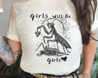 以下が含まれることがあります： クリーム色のTシャツ。黒と白のグラフィックで、ハートを持つカマキリが描かれ、「Girls will be Girls」の文字が入っています。デザインにはハートのシンボルと、昆虫の詳細なイラストが含まれています。