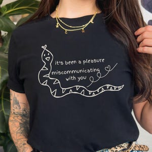 May include: Black t-shirt with a white snake illustration and the text "it's been a pleasure miscommunicating with you." The design includes a heart and a curved line. The shirt is made of a soft material.