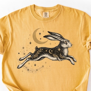 Puede incluir: Camiseta de color amarillo mostaza con una ilustración en blanco y negro de una liebre corriendo, una luna creciente y estrellas. El diseño incorpora elementos celestiales y botánicos. La camiseta tiene cuello redondo y mangas cortas.