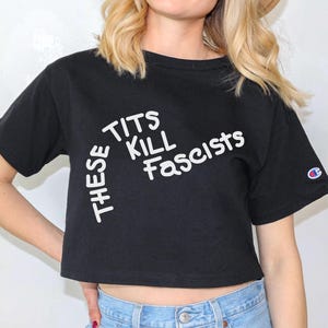 May include: Black crop top with the text "THESE TITS KILL Fascists" in white. The shirt has short sleeves and a Champion logo on the left sleeve.