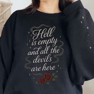 Puede incluir: Una sudadera negra de cuello redondo con el texto blanco "Hell is empty and all the devils are here" centrado en el pecho. El diseño incluye dos rosas de color rojo oscuro en la parte inferior y estrellas doradas en las mangas.