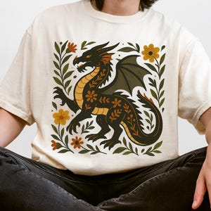 以下が含まれることがあります： クリーム色のTシャツ。黒いドラゴンのデザインに、黄色とオレンジのアクセント、花の要素が周りにあしらわれています。ドラゴンは翼を持ち、正方形の花柄のボーダーの中に配置されています。カジュアルウェアです。
