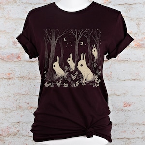 Op de afbeelding: Een maroon t-shirt met een afbeelding van vier witte konijnen in een bossetting. De konijnen kijken omhoog naar de maan, die wordt weergegeven door twee halve manen. De afbeelding is omringd door bomen en sterren.