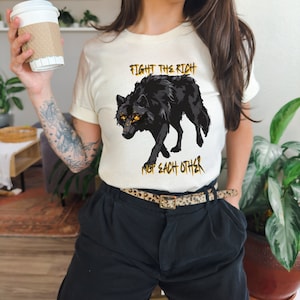 May include: Cream-colored t-shirt featuring a black wolf graphic with yellow eyes. The text "FIGHT THE RICH NOT EACH OTHER" is printed above and below the wolf in gold lettering. The shirt is paired with black pants and a leopard print belt.